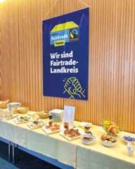 Dieser Alternativtext wurde KI-generiert: Ein blaues Banner mit Aufschrift hängt an einer Wand über einem gedeckten Tisch mit Speisen. Im Hintergrund ist ein Buffet mit verschiedenen Häppchen und Obst zu sehen.