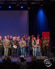 Dieser Alternativtext wurde KI-generiert: Eine Gruppe von Menschen steht auf einer Bühne vor einem Bühnenbild mit blauen Vorhängen. Einige halten Blumen oder Dokumente in den Händen.