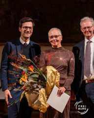 Dieser Alternativtext wurde KI-generiert: Drei Personen stehen vor einem dekorativen Hintergrund. Sie halten Blumen und ein weißes Blatt Papier.