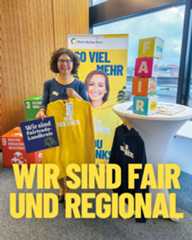 Dieser Alternativtext wurde KI-generiert: Eine Person steht vor einem Werbestand mit Bannern und Plakaten. Im Hintergrund ist ein Fenster mit Blick auf eine Stadt zu sehen.