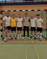 Dieser Alternativtext wurde KI-generiert: Eine Gruppe von Personen in Sportkleidung steht nebeneinander in einer Halle. Der Hintergrund besteht aus einer Wand mit bunten Feldern und einem Tor.