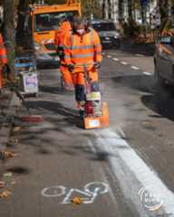 Dieser Alternativtext wurde KI-generiert: Auf einer Straße arbeiten Personen in orangefarbener Arbeitskleidung mit einer Maschine an der Fahrbahn. Im Hintergrund sind Fahrzeuge und Bäume zu sehen.
