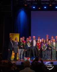 Dieser Alternativtext wurde KI-generiert: Eine Gruppe von Menschen steht auf einer Bühne vor blauen Vorhängen. Einige halten Blumen und stehen um eine Dekoration mit einem Geschenk herum.