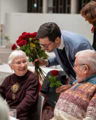 Dieser Alternativtext wurde KI-generiert: Eine Person überreicht Blumen an zwei ältere Menschen, die an einem Tisch sitzen. Im Hintergrund sind weitere Personen und eine helle, moderne Umgebung zu sehen.