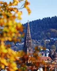 Dieser Alternativtext wurde KI-generiert: Ein hoher, spitzer Turm ragt über einer Stadtlandschaft mit vielen Gebäuden und umgebenden Hügeln auf. Herbstliche Blätter im Vordergrund verschwimmen mit der Szenerie.