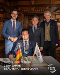 Dieser Alternativtext wurde KI-generiert: Schwarz-weißes Foto von mehreren Personen vor einer verzierten Holzwand. Im Vordergrund stehen Flaggen von Deutschland und Südkorea vor einem Tisch mit Aufschrift.