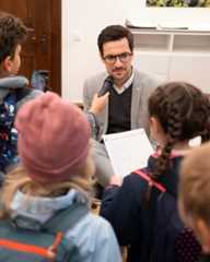 Dieser Alternativtext wurde KI-generiert: Eine Person mit Mikrofon spricht vor einer Gruppe von Kindern, die im Halbkreis sitzen und zuhören. Im Hintergrund befindet sich ein Regal und ein Fenster.