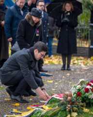 Dieser Alternativtext wurde KI-generiert: Mehrere Personen stehen oder knien vor einem Blumenschmuck. Eine Person legt ein Band oder eine Schleife zwischen die Blumen.