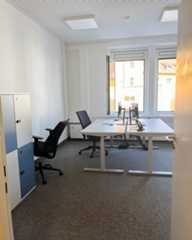 Dieser Alternativtext wurde KI-generiert: Ein heller Raum mit Büromöbeln, darunter ein Schreibtisch und Bürostühle. Durch das Fenster ist ein Blick auf Gebäude im Außenbereich erkennbar.