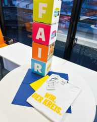 Dieser Alternativtext wurde KI-generiert: Ein Stapel farbiger Kartons mit den Buchstaben 'FAIR' steht auf einem Tisch. Daneben liegt ein blau-weißes Textil mit Aufdruck.