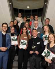 Dieser Alternativtext wurde KI-generiert: Eine Gruppe von Menschen steht auf einer Treppe und hält Dokumente oder Auszeichnungen in den Händen. Viele tragen Namensschilder mit einem grünen Logo.