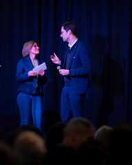 Dieser Alternativtext wurde KI-generiert: Zwei Personen stehen auf einer Bühne vor einem dunklen Hintergrund und sprechen vor einem Publikum. Beide halten Papier in den Händen.