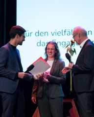 Dieser Alternativtext wurde KI-generiert: Eine Person erhält auf einer Bühne eine Auszeichnung und eine Blume von zwei weiteren Personen. Im Hintergrund befindet sich eine Projektion mit Text.