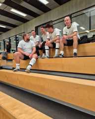 Dieser Alternativtext wurde KI-generiert: Mehrere Personen sitzen auf einer Bank in einem Umkleideraum. Sie tragen ähnliche Kleidung und scheinen sich zu unterhalten.