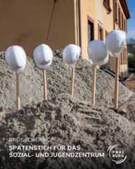 Dieser Alternativtext wurde KI-generiert: Mehrere weiße Schutzhelme stehen in einer Sandfläche, auf denen Schaufeln stecken. Im Bildtext ist von einem Spatenstich für ein Soziales- und Jugendzentrum die Rede.