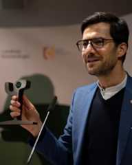 Dieser Alternativtext wurde KI-generiert: Eine Person mit Brille hält ein Mikrofon und spricht. Im Hintergrund sind weitere Personen und Logos zu sehen.