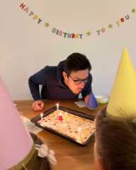 Dieser Alternativtext wurde KI-generiert: Eine Person pustet Kerzen auf einer Kuchenplatte aus. Im Hintergrund sind Dekorationen und weitere Personen zu sehen.