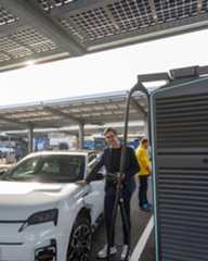 Dieser Alternativtext wurde KI-generiert: Ein Auto wird an einer Ladestation mit Solardach aufgeladen. Im Hintergrund sind weitere Fahrzeuge und ein Gebäude erkennbar.