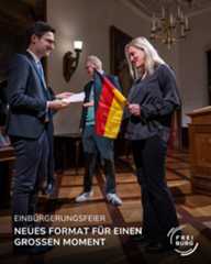 Dieser Alternativtext wurde KI-generiert: Eine Aufnahme zeigt Menschen in einem Innenraum während einer Zeremonie. Eine Person überreicht einer anderen ein Dokument, während im Hintergrund eine Flagge zu sehen ist.