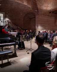 Dieser Alternativtext wurde KI-generiert: Ein Innenraum mit einer gewölbten, roten Backsteinwand. Menschen sitzen und stehen vor einer Bühne mit einem Flügel und beobachten eine Aufführung.