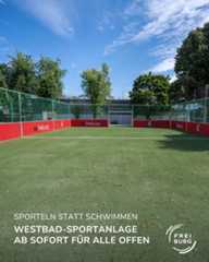 Dieser Alternativtext wurde KI-generiert: Ein Sportplatz mit grünem Rasen und rotem Zaun ist zu sehen. Im Vordergrund steht ein Plakat mit Text und einem Logo.