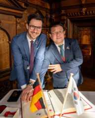 Dieser Alternativtext wurde KI-generiert: Zwei Personen in Anzügen stehen nebeneinander vor einem großen, dunklen Holztisch. Auf dem Tisch befinden sich Flaggen sowie ein Smartphone und ein geöffnetes Buch.