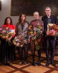 Dieser Alternativtext wurde KI-generiert: Vier Personen stehen nebeneinander und halten Blumensträuße. Das Foto ist farbig und zeigt eine Gruppe vor einer dunklen Wand mit einem Gemälde.