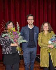 Dieser Alternativtext wurde KI-generiert: Drei Personen stehen auf einer Bühne vor roten Vorhängen und halten Blumensträuße. Das Bild ist farbig und zeigt eine Nahaufnahme der Personen.