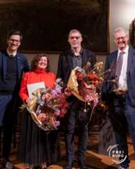 Dieser Alternativtext wurde KI-generiert: Ein farbiges Porträt zeigt drei Personen und einen großen Blumenstrauß vor einem dunklen Hintergrund mit einem Bild an der Wand. Die Personen stehen nebeneinander und lächeln in die Kamera.