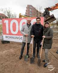 Dieser Alternativtext wurde KI-generiert: Drei Personen stehen lächelnd mit Schaufeln vor einer Baustelle und einer Werbetafel. Im Hintergrund ist ein Gebäude und ein Bagger zu sehen.