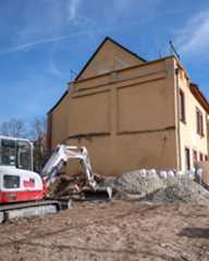 Dieser Alternativtext wurde KI-generiert: Ein Gebäude mit einem hellen Putz steht neben einer Baustelle. Im Vordergrund ist ein Bagger zu sehen, der auf einem unbefestigten Grundstück steht.