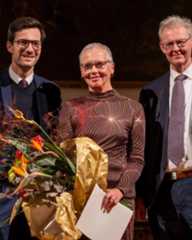 Dieser Alternativtext wurde KI-generiert: Drei Personen stehen auf einer Bühne vor einem Hintergrund mit Paneelen. Eine Blumenkomposition und ein golden umwickeltes Geschenk befinden sich im Vordergrund.