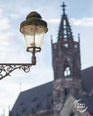 Dieser Alternativtext wurde KI-generiert: Eine historische Straßenlaterne im Vordergrund, mit einem spitzen Kirchturm im Hintergrund unter einem blauen Himmel. Das Bild zeigt eine Stadtansicht.