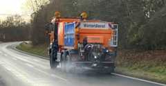 Dieser Alternativtext wurde KI-generiert: Ein orangefarbenes Fahrzeug mit Aufschrift "Winterdienst" fährt auf einer Straße. Im Hintergrund ist eine Landschaft mit Bäumen und einer Straße zu sehen.