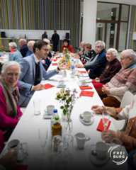 Dieser Alternativtext wurde KI-generiert: Eine Gruppe von Menschen sitzt an einem langen Tisch und unterhält sich bei einer Mahlzeit. Auf dem Tisch stehen Getränke, Geschirr und Blumen.