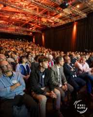 Dieser Alternativtext wurde KI-generiert: Ein Publikum sitzt in einem großen, hell beleuchteten Raum mit einer Bühne. Die Beleuchtung ist überwiegend rot und orange, was eine warme Atmosphäre erzeugt.