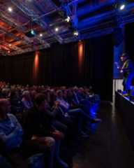 Dieser Alternativtext wurde KI-generiert: Eine große Menschenmenge sitzt in einem dunklen Raum und blickt auf eine Bühne. Das Bild ist in Blau- und Rottönen gehalten und zeigt eine weitläufige Hallenstruktur.