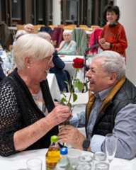 Dieser Alternativtext wurde KI-generiert: Zwei Personen sitzen an einem gedeckten Tisch und lächeln sich an. Eine Person überreicht eine einzelne rote Rose.