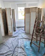 Dieser Alternativtext wurde KI-generiert: Ein Raum mit mehreren großen, verpackten Gegenständen und einer Leiter. Der Boden ist mit Folie abgedeckt, und im Hintergrund ist eine Tür zu sehen.