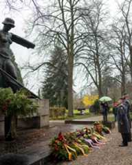 Dieser Alternativtext wurde KI-generiert: Eine Aufnahme zeigt ein Denkmal im Freien, dekoriert mit Blumen und Tannenzweigen. Im Hintergrund stehen Personen unter einem Baum, einige halten Regenschirme.