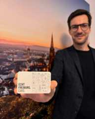 Dieser Alternativtext wurde KI-generiert: Eine Person hält eine Karte vor einer Stadtlandschaft bei Sonnenuntergang in die Kamera. Die Karte zeigt verschiedene Symbole und den Text