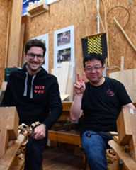 Dieser Alternativtext wurde KI-generiert: Zwei Personen sitzen nebeneinander auf einer Holzbank in einem Werkraum. Im Hintergrund sind Werkzeuge und Holzstücke sowie ein gerahmtes Bild zu sehen.