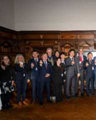 Dieser Alternativtext wurde KI-generiert: Eine Gruppe von Menschen steht in einem Raum mit dekorativen Holztäfelungen. Mehrere Personen zeigen mit ihren Händen ein Herzsymbol.