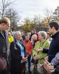 Dieser Alternativtext wurde KI-generiert: Eine Gruppe von Menschen steht und unterhält sich im Freien. Im Hintergrund sind Bäume mit Herbstlaub und Gebäude zu sehen.