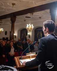 Dieser Alternativtext wurde KI-generiert: Ein Innenraum mit festlicher Dekoration, darunter Kronleuchter und Stuck. Eine Person steht mit dem Rücken zum Betrachter an einem Rednerpult vor einer Gruppe von Menschen.