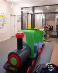 Dieser Alternativtext wurde KI-generiert: Ein Spielzeugzug steht im Vordergrund, vor einer Glastür zu einem Raum mit Spielzeug und Dekoration an der Wand. Das Bild ist farbig und zeigt eine helle, freundliche Umgebung.