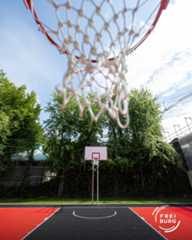 Dieser Alternativtext wurde KI-generiert: Ein Basketballkorb steht auf einem roten Basketballfeld. Im Hintergrund sind Bäume und Gebäude unter blauem Himmel zu sehen.