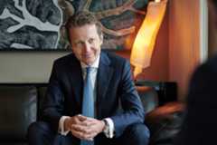 Renaud de Planta, associé principal de Pictet, assis sur un canapé, vêtu d’un costume sombre et cravate bleue, avec une lampe moderne en arrière-plan.