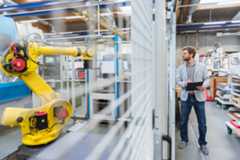 Un homme en veste grise observe un robot industriel jaune dans une usine moderne.