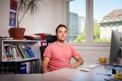 Un homme assis à un bureau dans un bureau lumineux à Lausanne, avec des étagères remplies de dossiers et une plante en pot à l’arrière-plan.